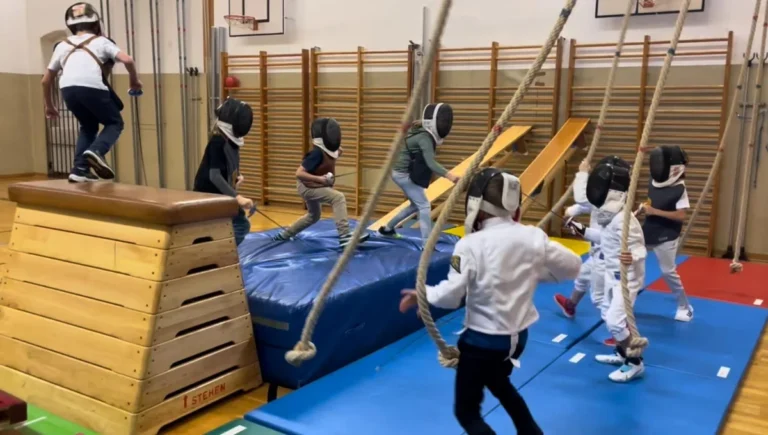 Kinder mit Fechtmasken und Übungswaffen spielen in einer Turnhalle auf Matten und an Turngeräten.
